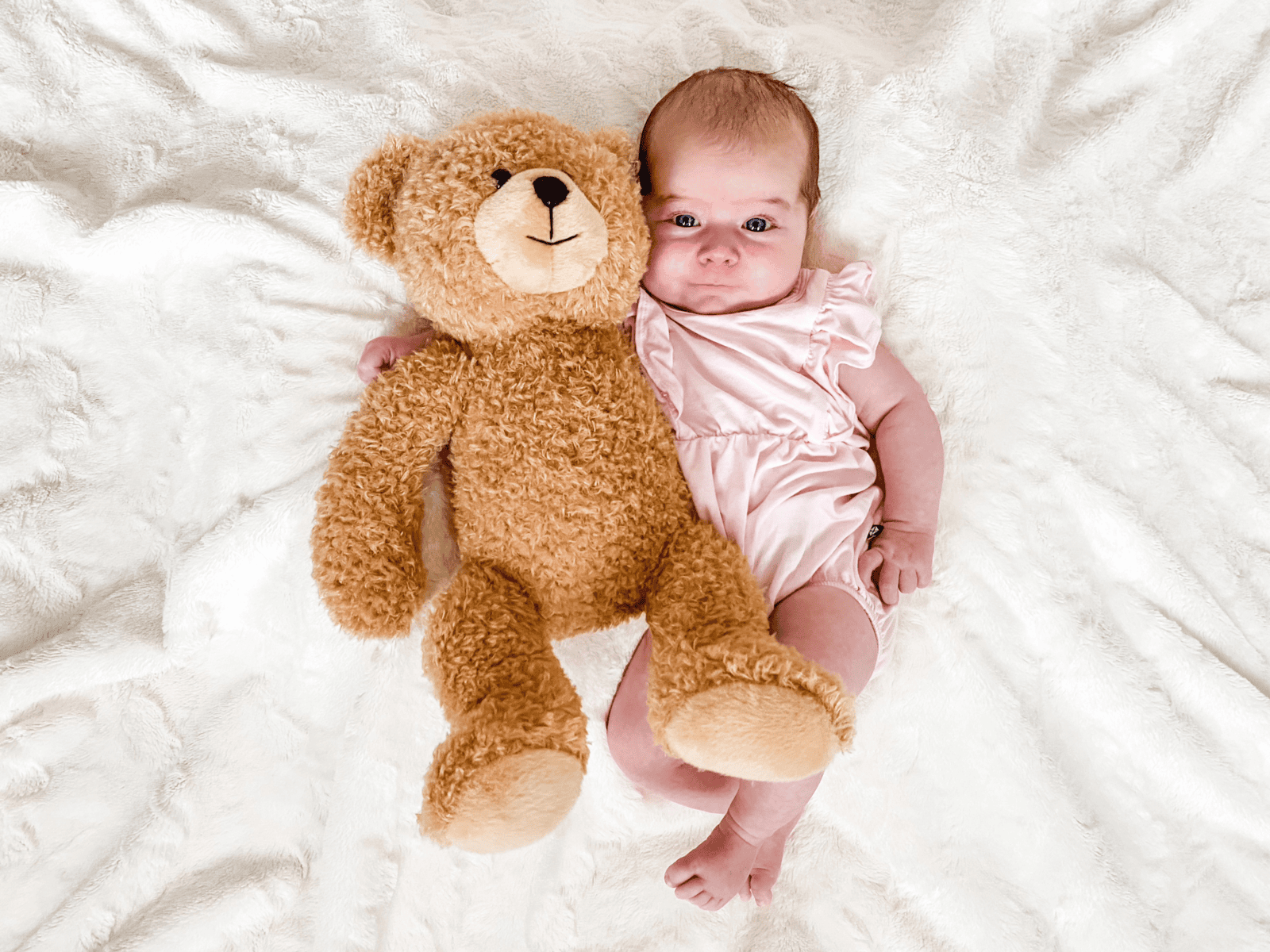 Two-month-old baby lying on a white blanket in a light pink romper beside a teddy bear, looking up with wide blue eyes.
