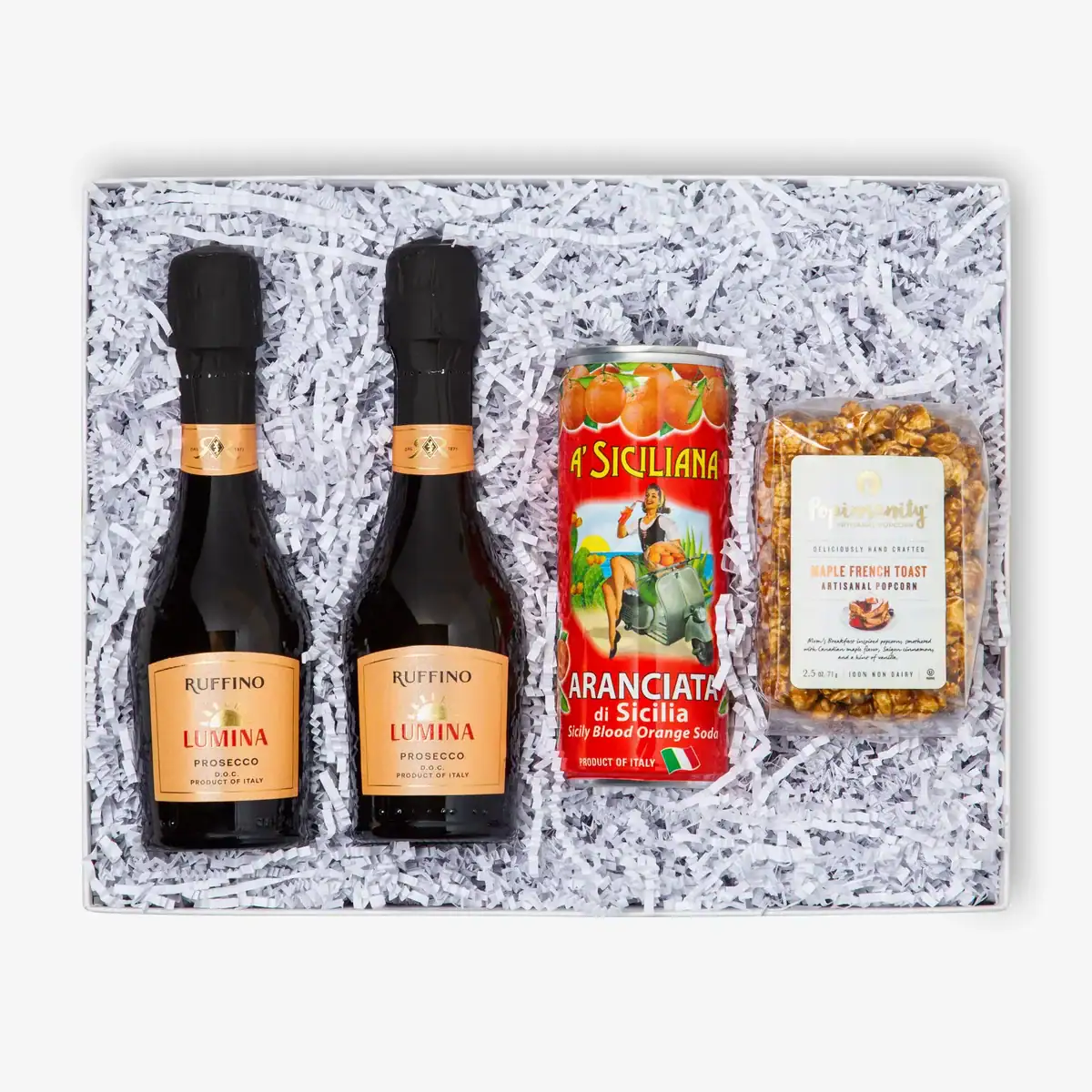 A baby shower mimosa party favor kit featuring two mini bottles of Ruffino Lumina Prosecco, a can of Sicilian blood orange soda, and a bag of maple french toast artisanal popcorn, all neatly arranged in a white gift box with shredded filler.