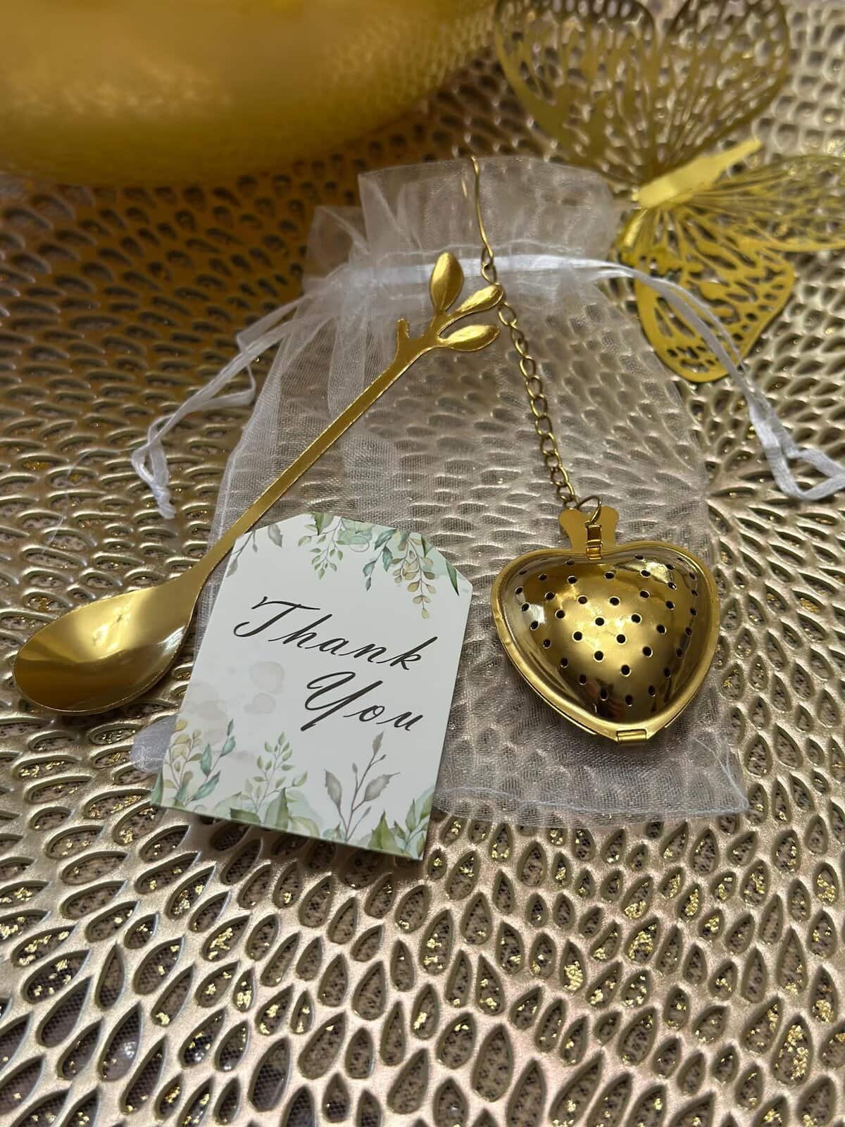 Elegant baby shower party favor featuring a gold heart-shaped tea infuser and matching gold spoon with a leaf handle, displayed on a white mesh bag with a botanical “Thank You” tag. Set on a textured gold background with a decorative gold butterfly