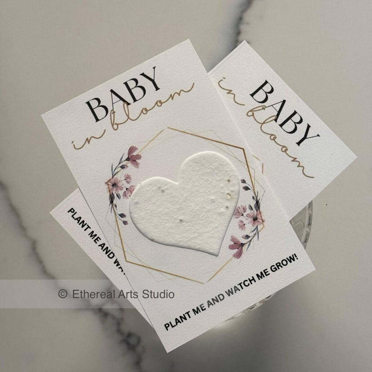Elegant baby shower party favors featuring seed paper cards with a heart-shaped biodegradable insert. The cards read “Baby in Bloom” with floral accents and the message “Plant me and watch me grow!” — perfect for a garden-themed baby shower.