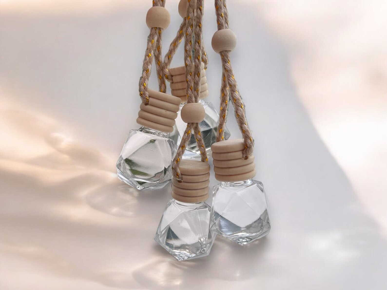 Clear faceted glass diffuser bottles filled with liquid and topped with light wooden caps and beaded rope hangers, displayed against a soft neutral background with gentle sunlight reflections.