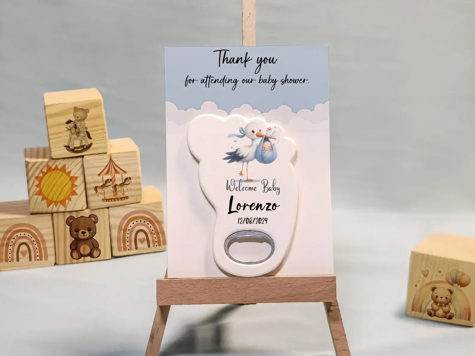 Baby shower favor featuring a stork design bottle opener on a personalized card with the name “Lorenzo” and a baby shower date. The card reads “Thank you for attending our baby shower” and is displayed on a small wooden easel, surrounded by wooden toy blocks with playful designs like rainbows, teddy bears, and a carousel.