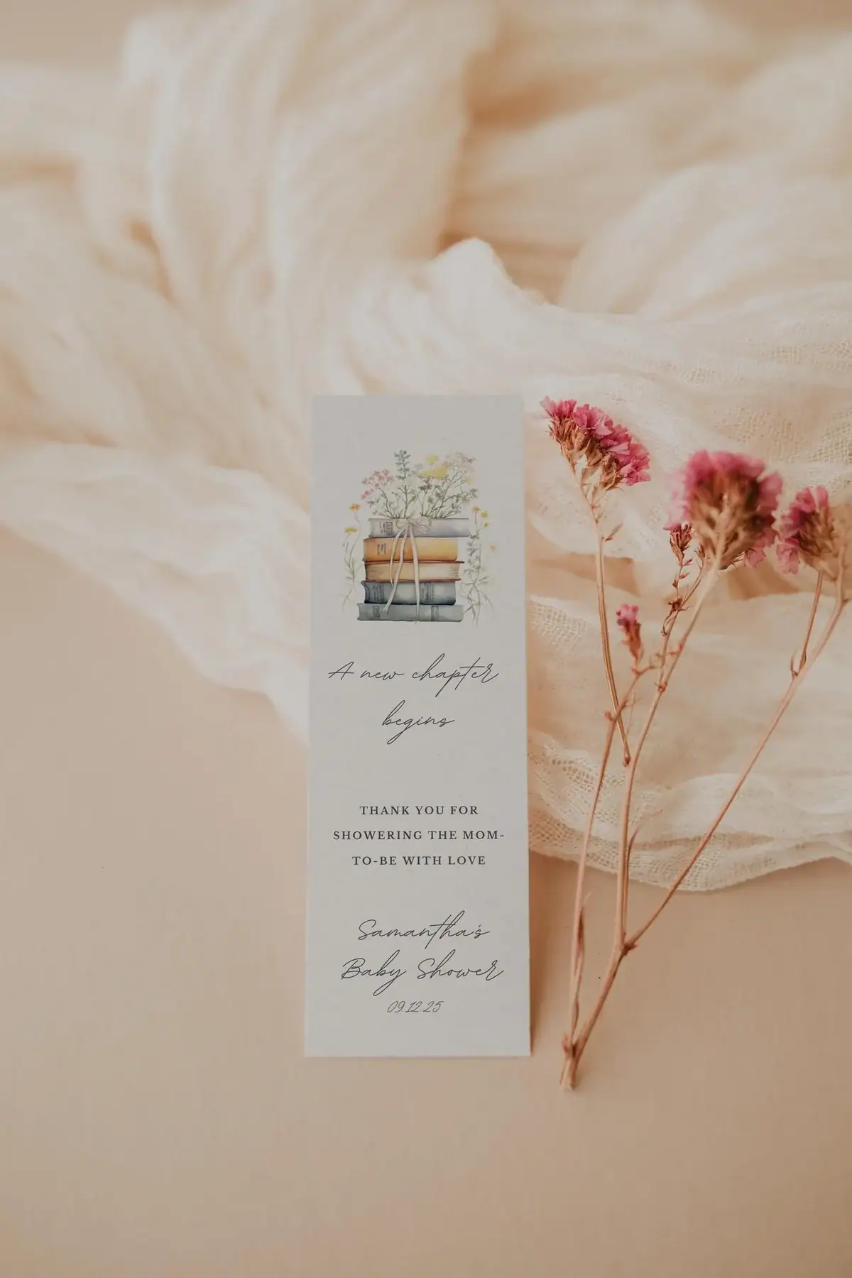 Minimalist baby shower bookmark favor featuring a stack of books with floral accents and soft script text, styled against a blush background with delicate pink flowers and a light textured fabric for a soft, romantic aesthetic.