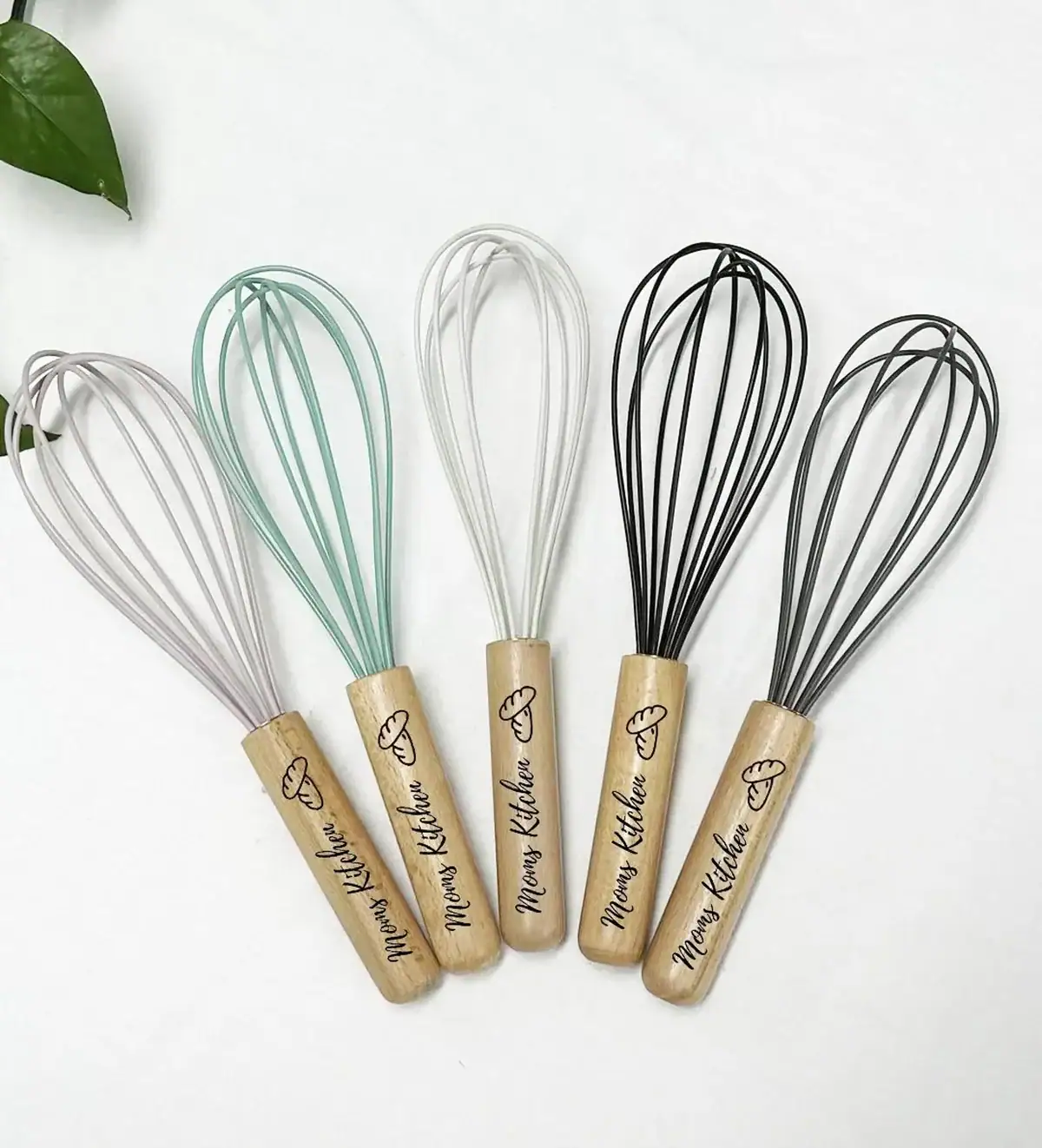 Flat lay of five silicone kitchen whisks in assorted pastel and neutral colors with wooden handles engraved with the words “Mom’s Kitchen,” placed on a white background with a green leaf in the corner.