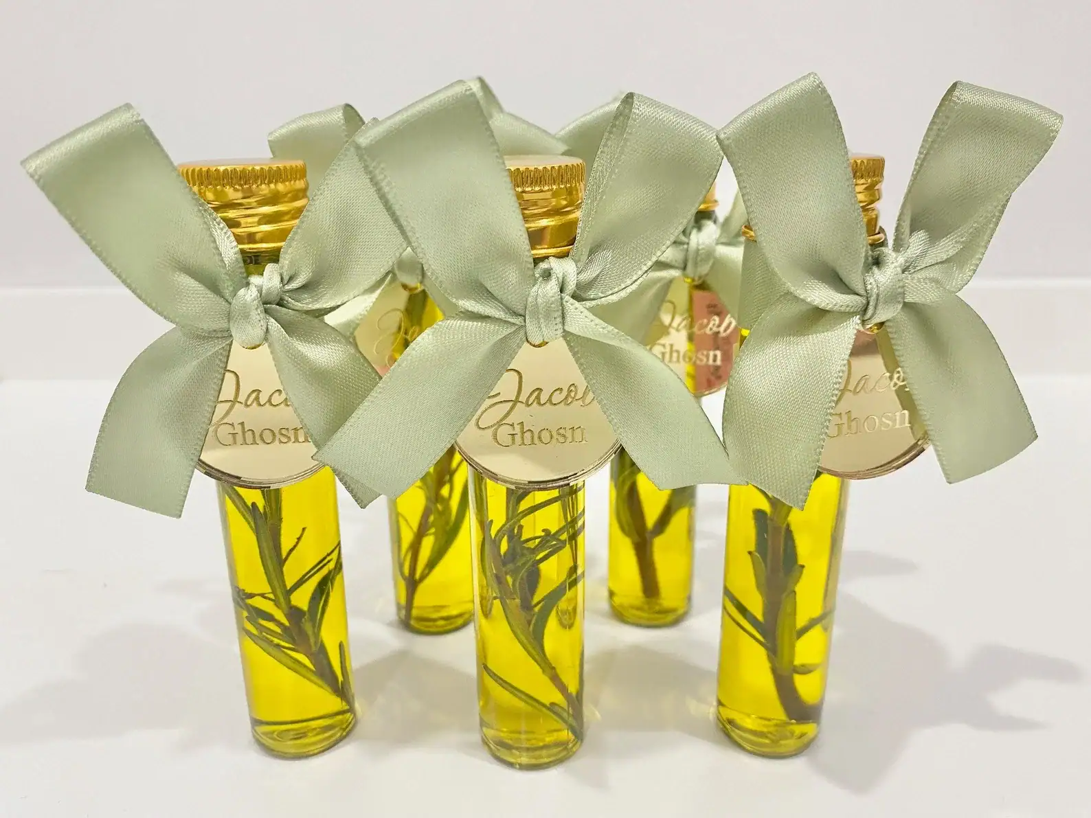 Close-up of mini glass bottles filled with olive oil and sprigs of rosemary, each sealed with a gold cap and decorated with a sage green satin bow and personalized gold tag, arranged neatly in rows.