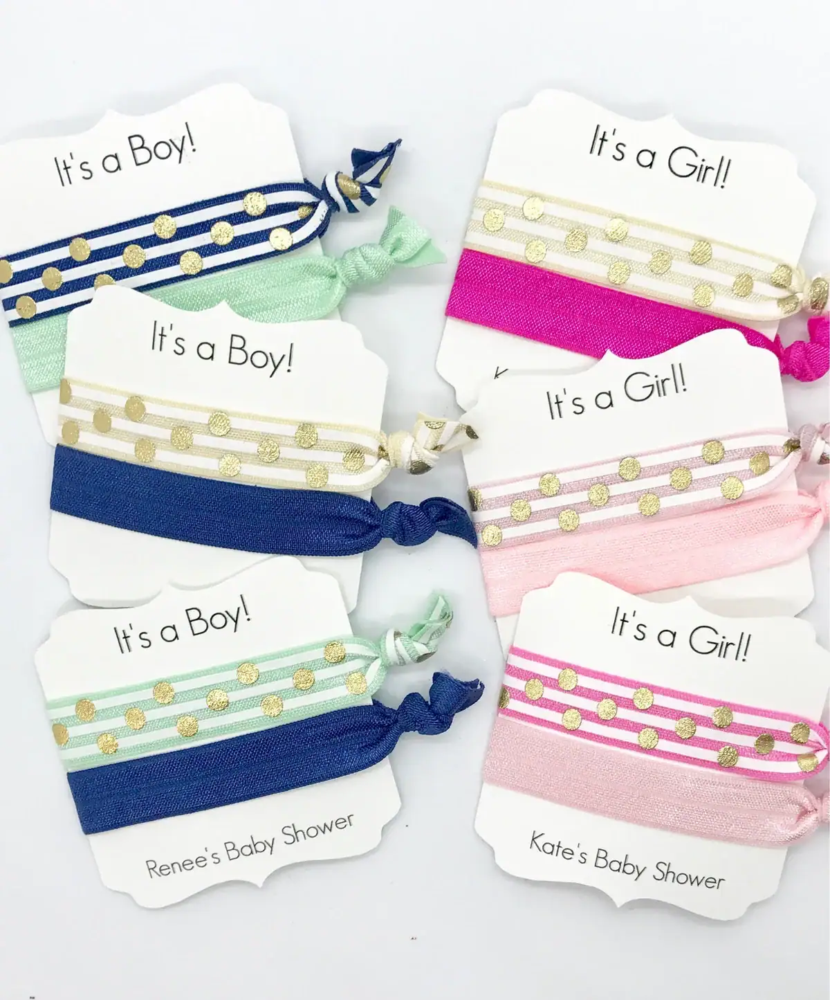 Flat lay of baby shower hair tie favors labeled “It’s a Boy!” and “It’s a Girl!” featuring stretchy bands in navy, mint green, hot pink, and blush with gold polka dot accents, displayed on white cards.