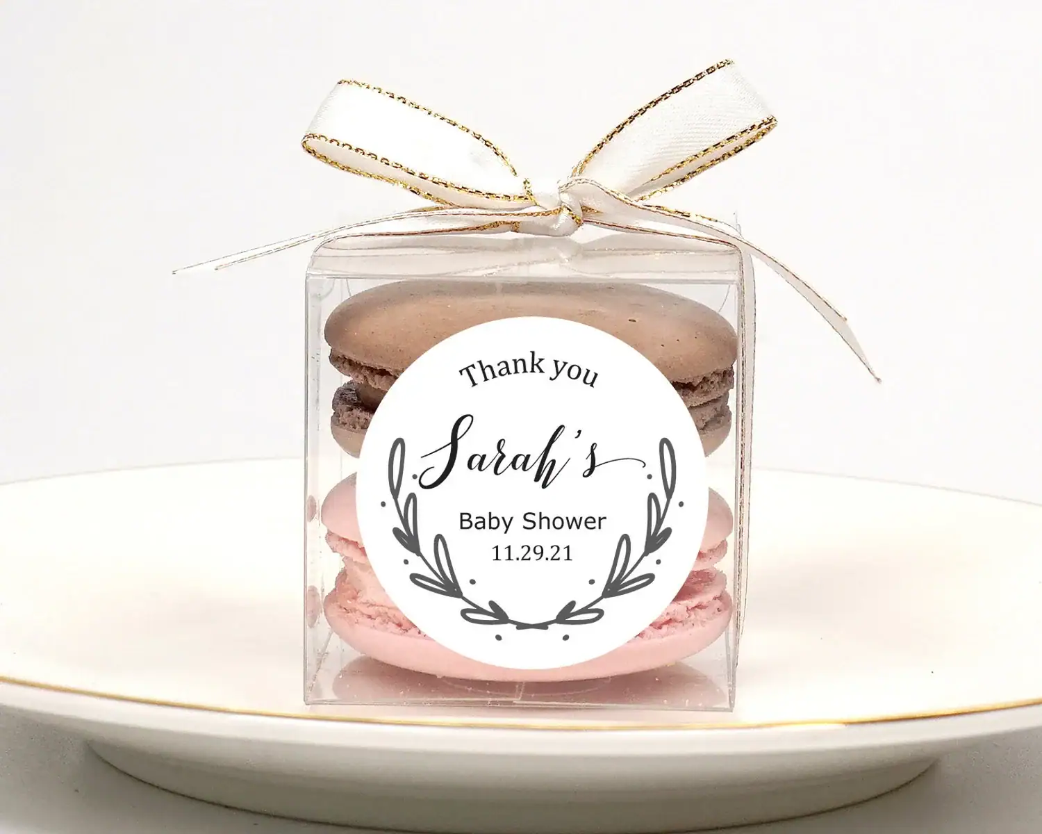 Clear plastic baby shower party favor box filled with pink and chocolate macarons, tied with a white ribbon with gold trim, and labeled with a personalized sticker reading “Thank you, Sarah’s Baby Shower, 11.29.21.”