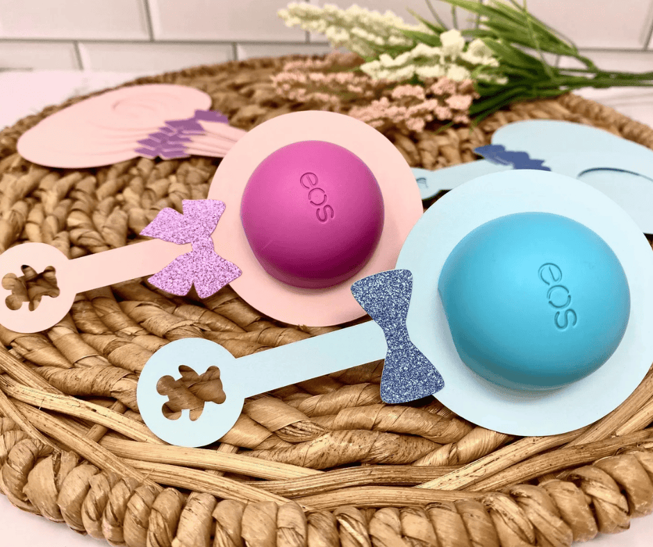 Close-up of EOS lip balm party favors shaped like baby rattles, featuring pink and blue pastel designs with glittery bows, perfect for a baby shower.