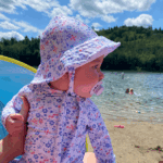 Baby wearing a floral sunhat and rash guard sitting on the beach with a pacifier, looking out at the lake on a sunny summer day.