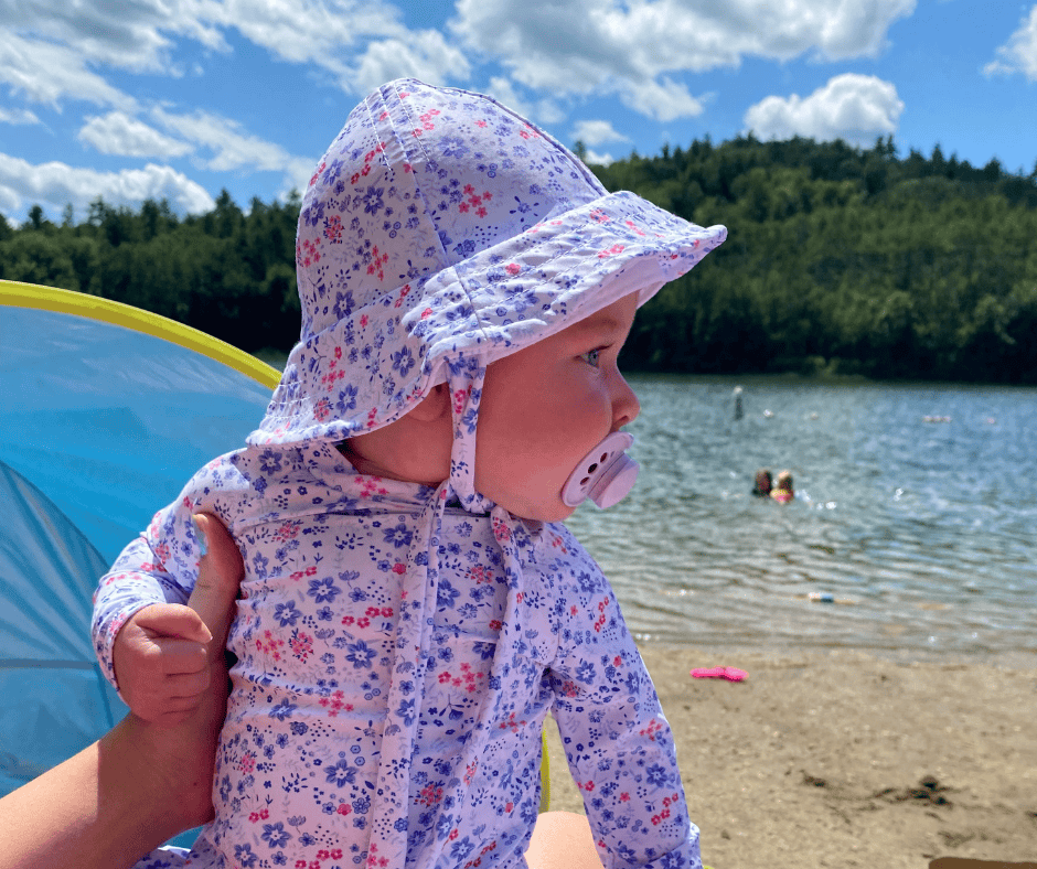 Baby wearing a floral sunhat and rash guard sitting on the beach with a pacifier, looking out at the lake on a sunny summer day.