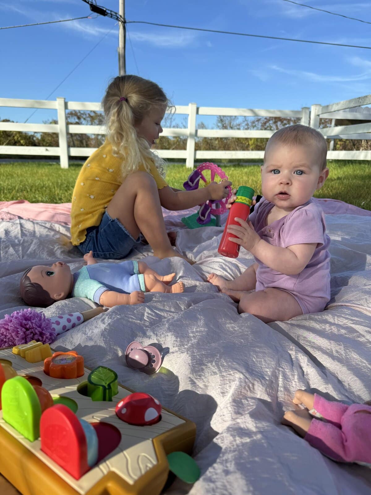 Two young children playing outside on a large beach blanket in the grass, surrounded by toys and sunshine, using the blanket as a soft play space in the backyard.