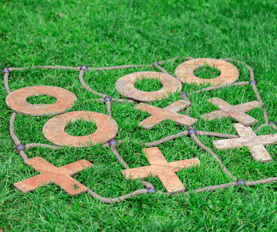 Outdoor tic-tac-toe game made with rustic wooden X and O pieces laid on a rope grid on green grass.