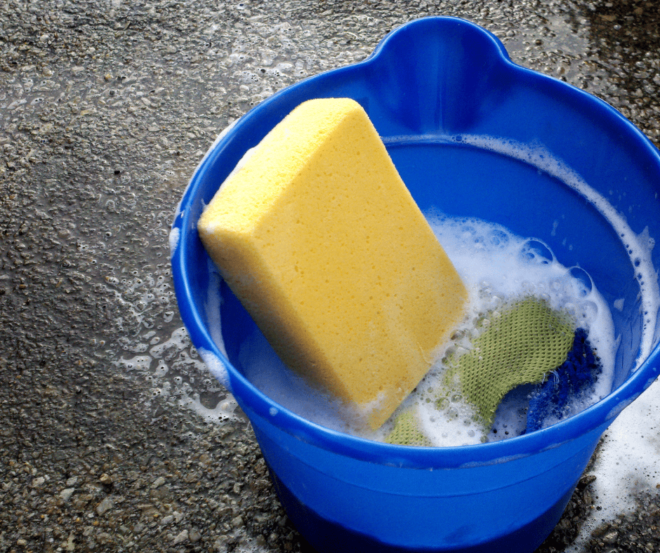 Yellow sponge resting in a blue plastic bucket filled with soapy water and cleaning cloths on a wet pavement.