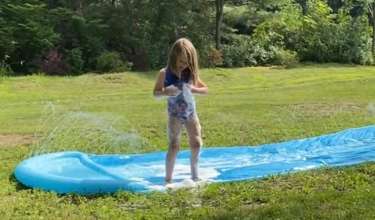 Child playing on a blue slip and slide in the backyard, covered in soapy bubbles as water sprays from the sides.