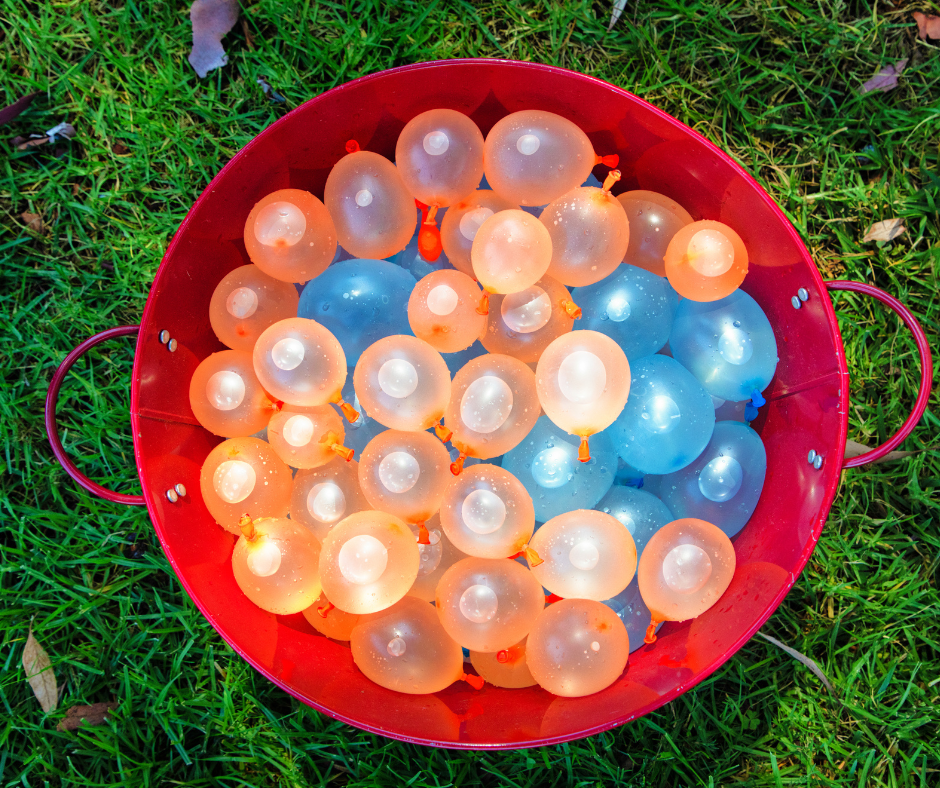 Top-down view of a red bucket filled with colorful orange and blue water balloons, ready for a backyard water fight on green grass.