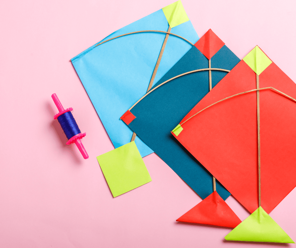 Three colorful homemade kites made from paper, string, and wooden sticks laid out on a pink background next to a spool of blue kite string.