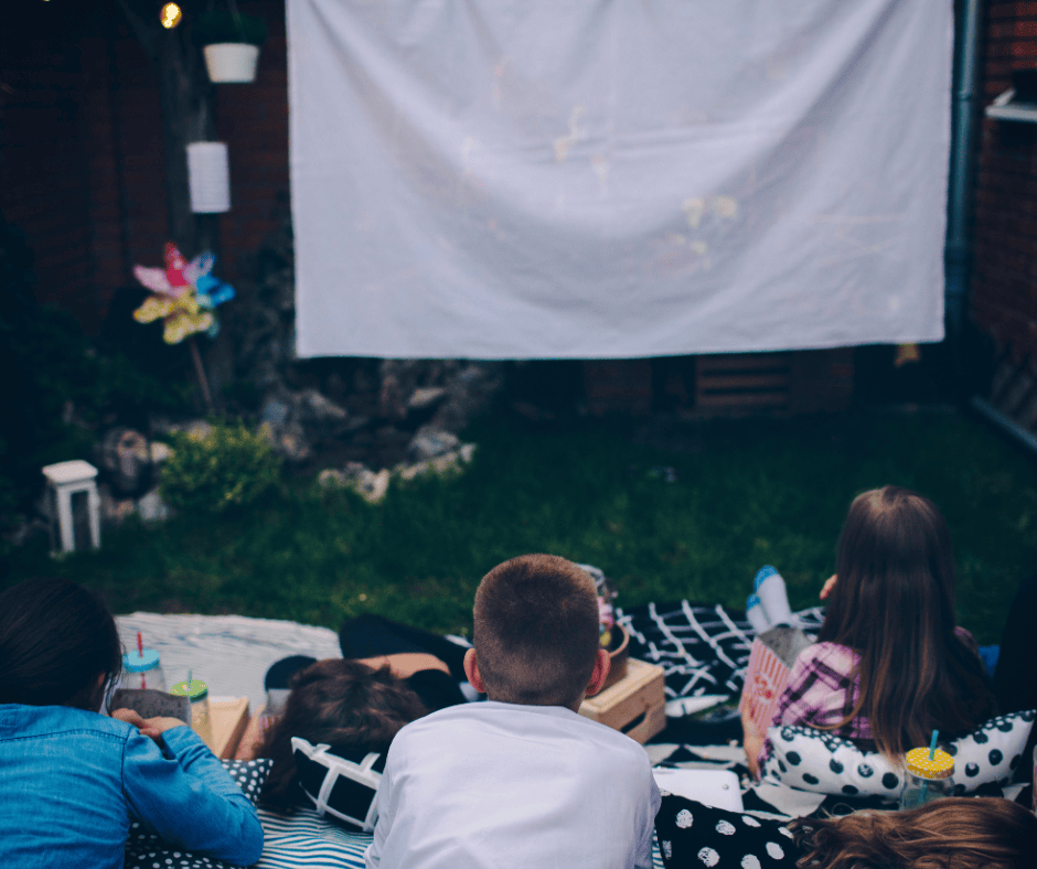 Children lying on blankets and pillows in the backyard, watching a movie projected onto a white sheet screen during an outdoor movie night.