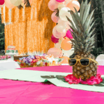 Luau-themed birthday party table decorated with a pineapple wearing yellow sunglasses, tropical leaves, and pink flowers, with a colorful balloon arch and fringe backdrop in the background.