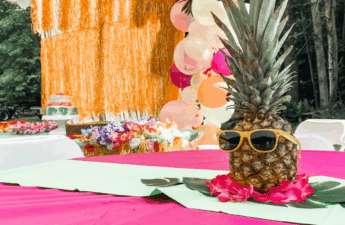 Luau-themed birthday party table decorated with a pineapple wearing yellow sunglasses, tropical leaves, and pink flowers, with a colorful balloon arch and fringe backdrop in the background.