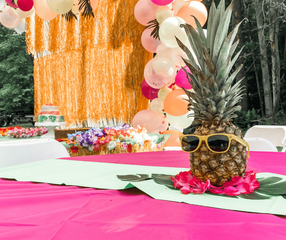 Luau-themed birthday party table decorated with a pineapple wearing yellow sunglasses, tropical leaves, and pink flowers, with a colorful balloon arch and fringe backdrop in the background.