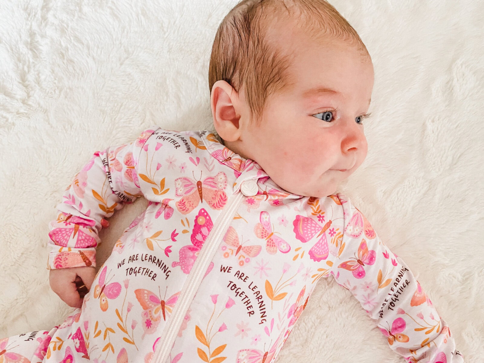 Newborn baby lying on a white fuzzy blanket wearing soft bamboo pajamas with a pink butterfly print and the phrase “We are learning together.”