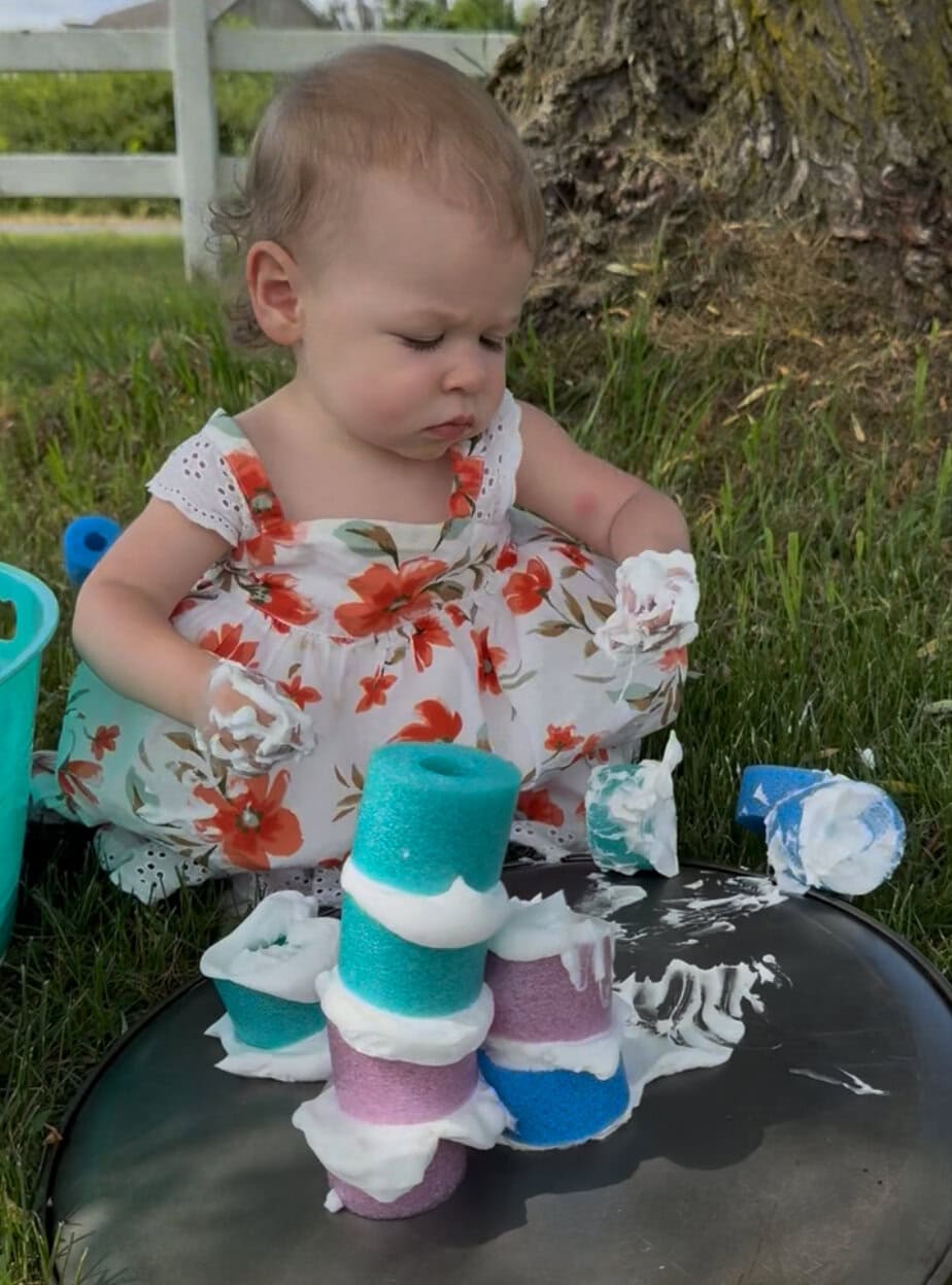 A toddler in a floral dress explores sensory play by stacking pieces of pool noodles with shaving cream in the grass under a tree.