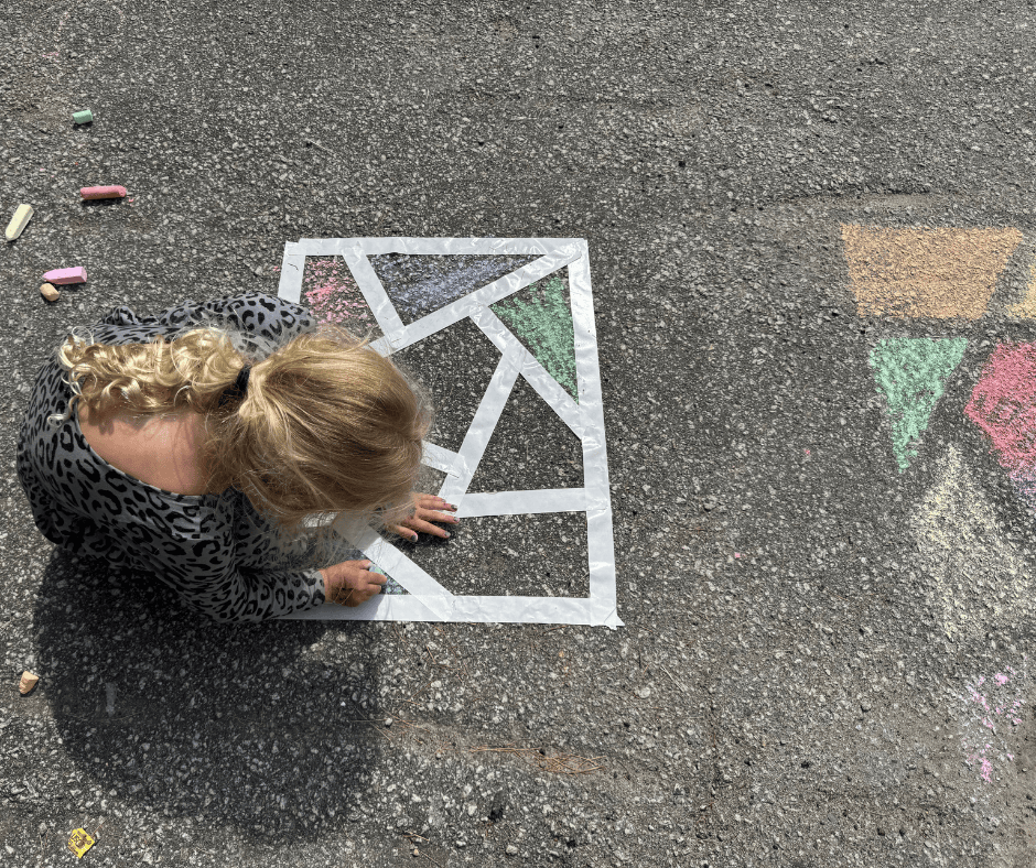 A young girl with blonde curls creates stained glass chalk art on the pavement using colorful sidewalk chalk and painter’s tape to form geometric shapes.