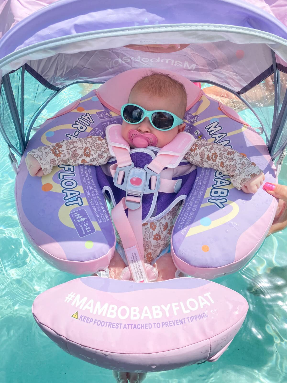Baby floating in a pool wearing sunglasses and a pacifier, secured in a pink and purple Mambo Baby Float with a canopy for sun protection.