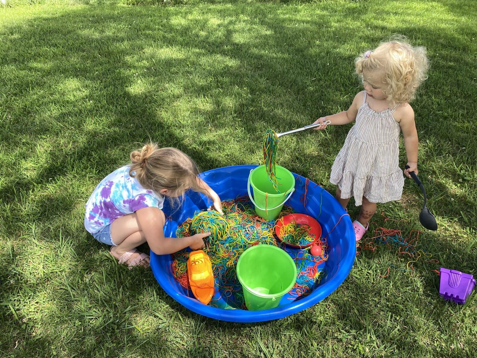 Child playing with rainbow-colored spaghetti in a kiddie pool outside for a fun summer activity for kids.