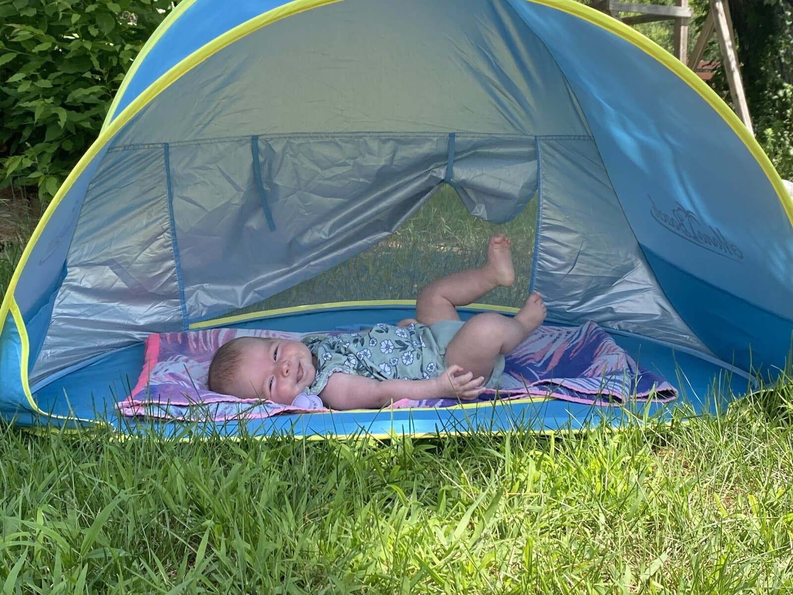 Baby lying on a towel inside a blue pop-up baby beach tent with mesh sides, smiling and relaxing in the shade on a sunny summer day.