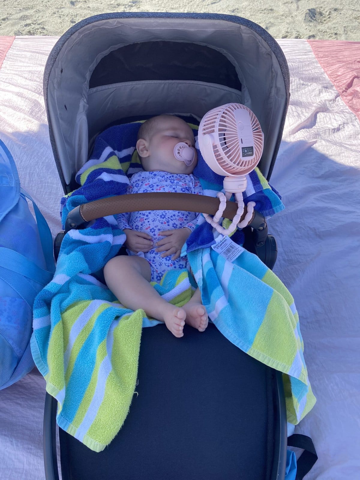 Baby sleeping in a stroller at the beach with a pink clip-on fan blowing on her face, wrapped in colorful beach towels for shade and comfort.