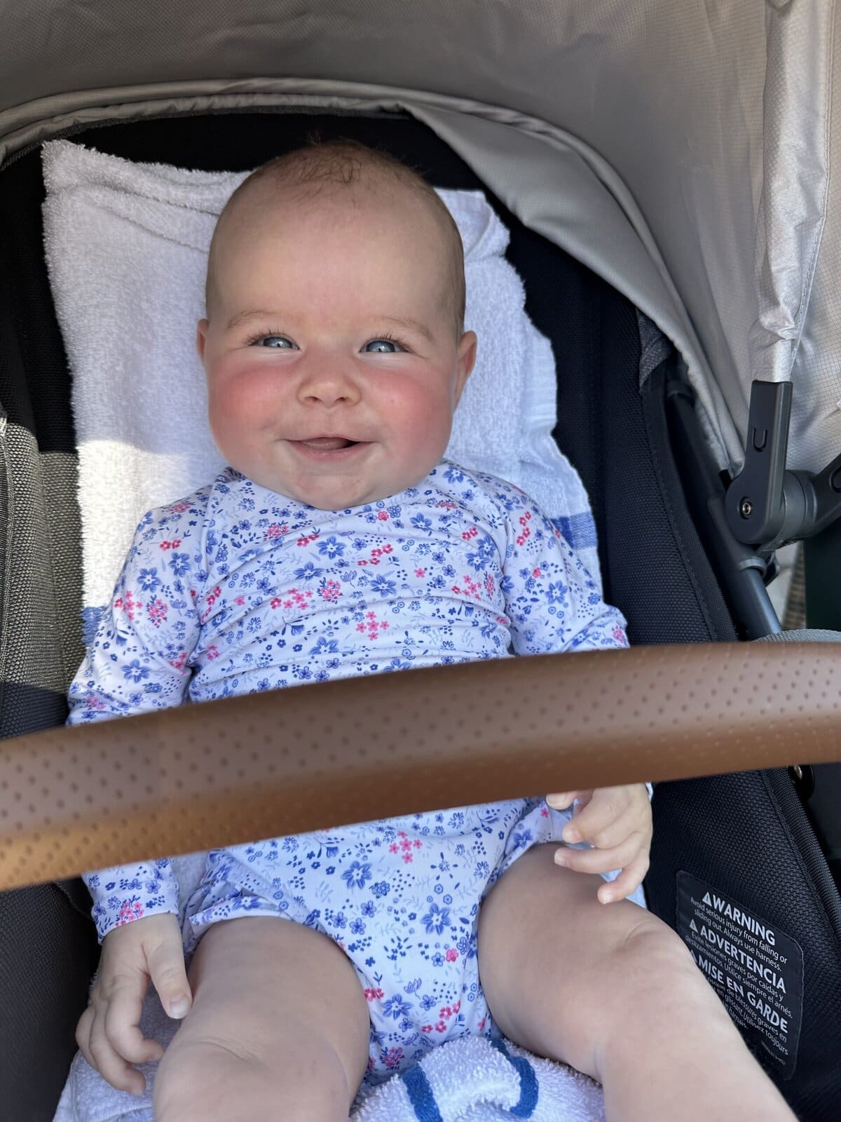 Smiling baby sitting in a stroller wearing a floral rash guard bathing suit, enjoying a sunny day outdoors with a white towel for comfort.