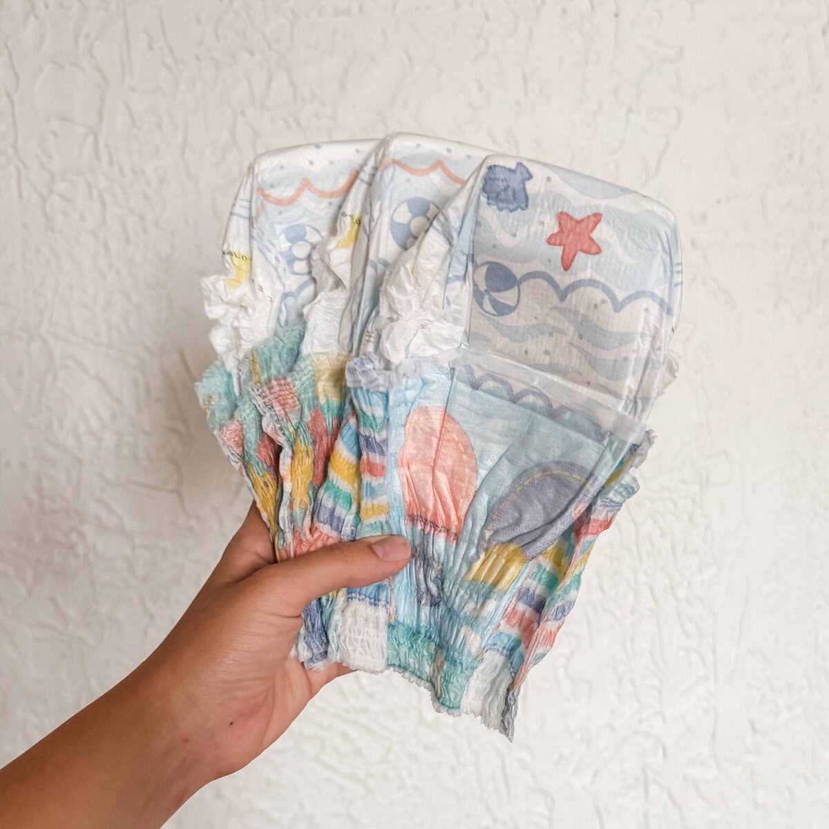 Hand holding a fan of colorful disposable swim diapers with beach-themed prints, perfect for summer pool and beach days with baby.