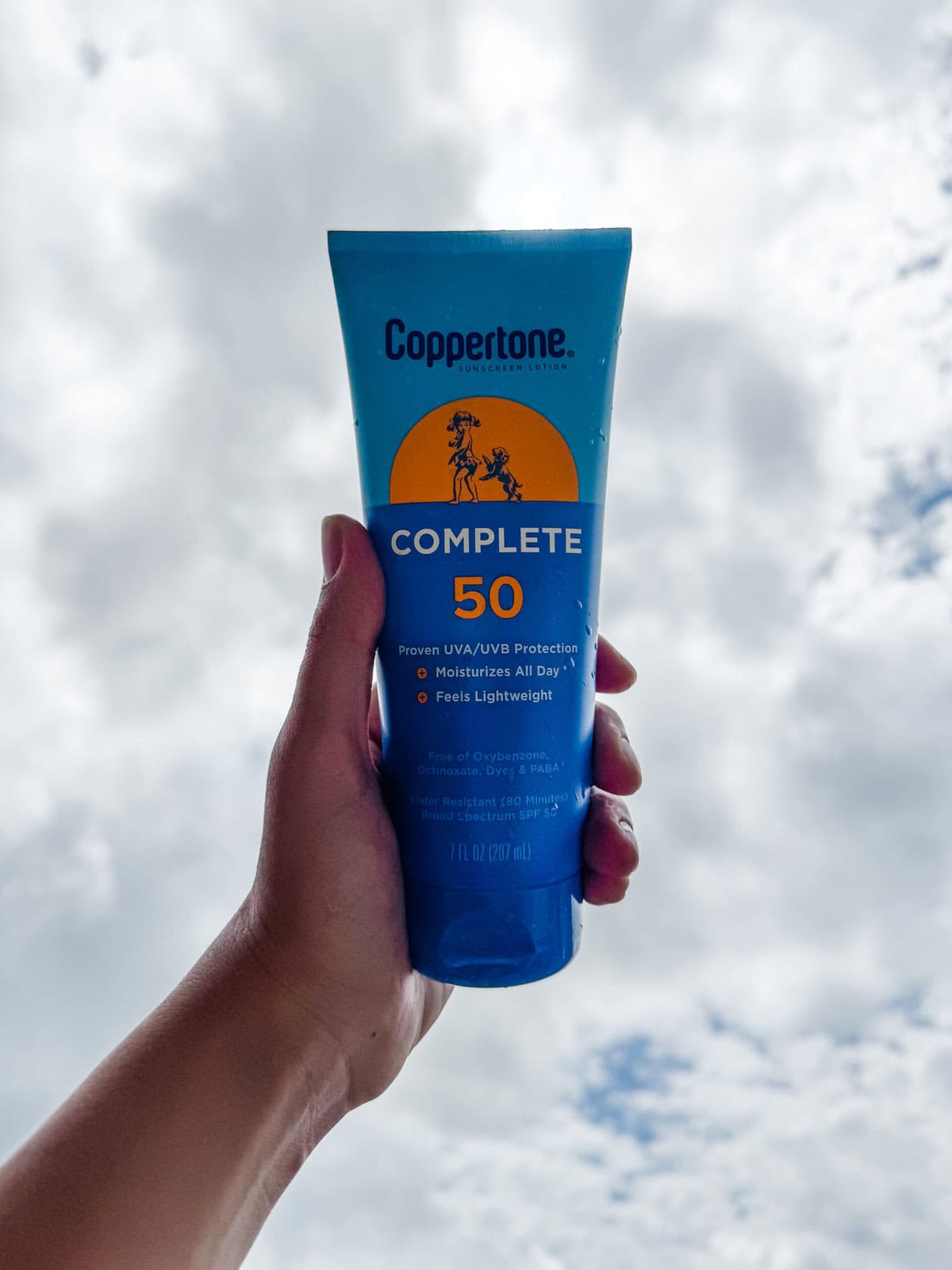Hand holding a bottle of Coppertone SPF 50 sunscreen lotion up to a cloudy summer sky, highlighting baby-safe sun protection for outdoor activities.