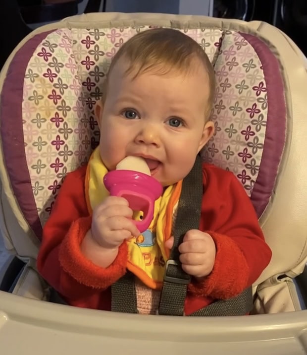 Baby sitting in a high chair enjoying a frozen breastmilk popsicle in a pink silicone mold, wearing a red outfit and colorful bib on a warm summer day.