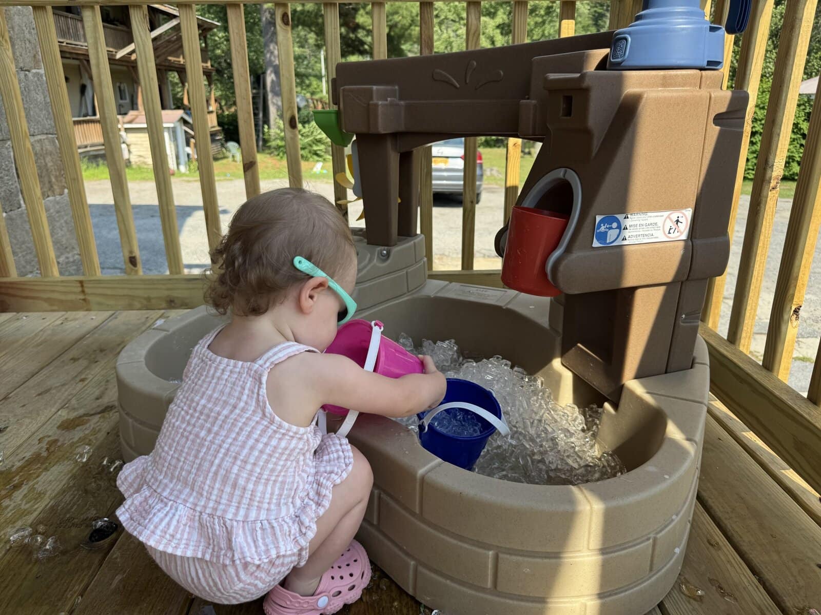 A toddler wearing a pink gingham romper and green sunglasses plays with ice and buckets at a water table on a wooden deck during a summer afternoon.