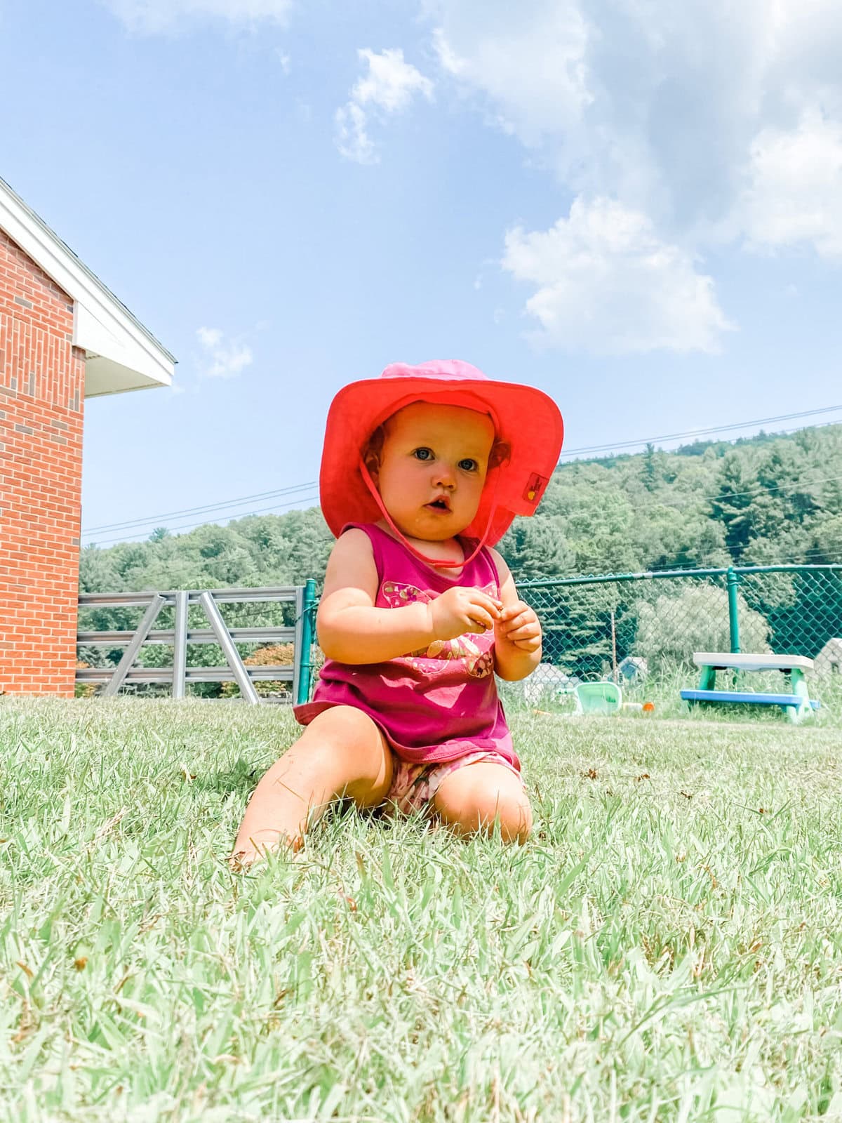 Toddler sitting in the grass on a sunny day wearing a bright pink sunhat and summer outfit, protected from the sun while playing outside.