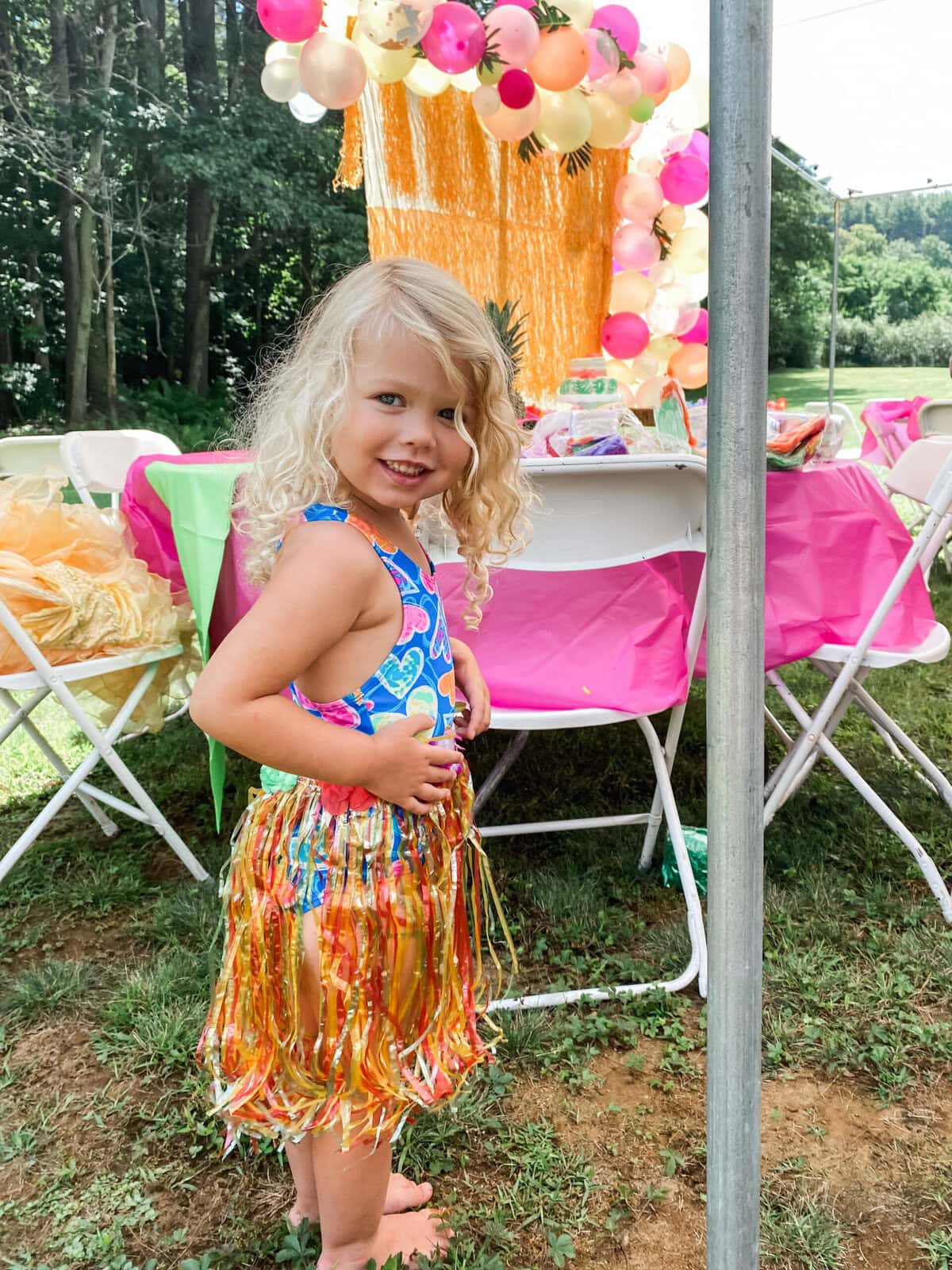 Young girl smiling at a luau-themed birthday party, wearing a colorful heart-print swimsuit and shiny grass skirt, with festive balloon decorations and pink table settings in the background.