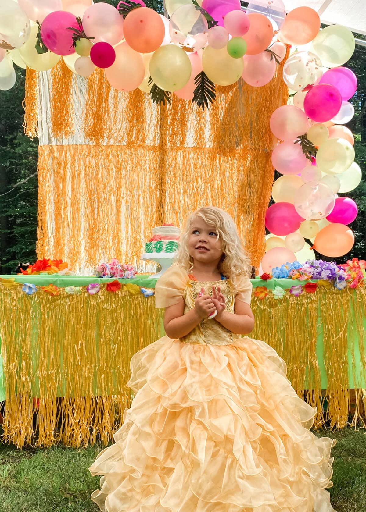 Young girl in a yellow princess dress smiling in front of a luau-themed cake table decorated with colorful leis, a grass skirt table cover, and a balloon arch with tropical leaves.