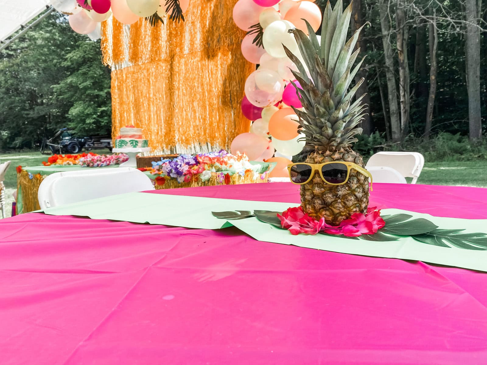 Tropical luau party table decorated with a pink tablecloth, green leaf runner, and a pineapple wearing yellow sunglasses and a flower lei, with a festive balloon and fringe backdrop in the background.