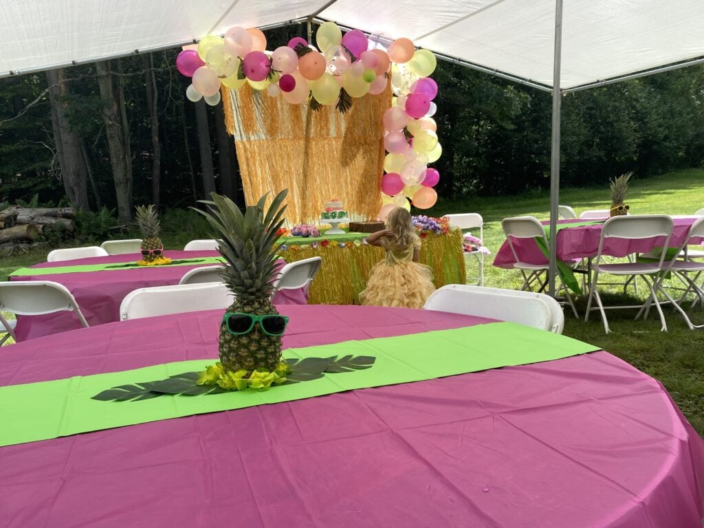 Outdoor luau birthday party setup with pink and green tables, pineapples wearing sunglasses as centerpieces, and a balloon and fringe backdrop where a young girl in a yellow dress stands near the cake table.