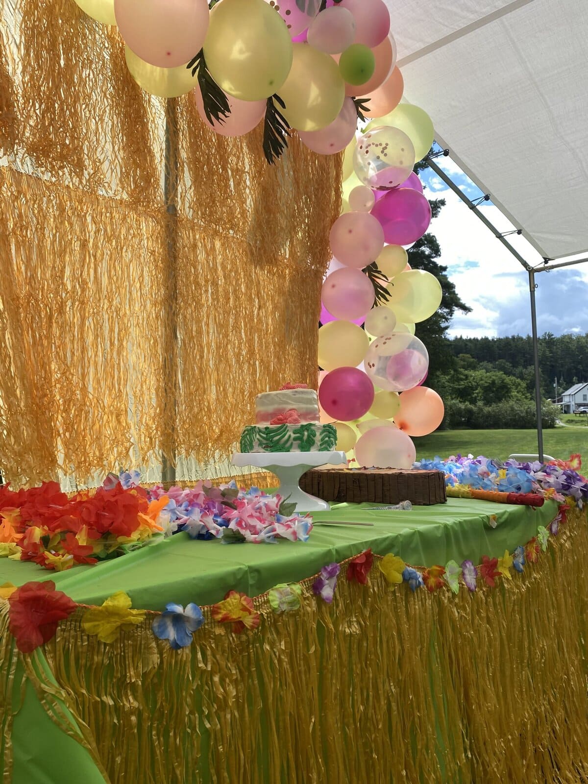 Luau-themed cake table decorated with a green tablecloth, yellow fringe skirt, colorful leis, and a two-tier tropical cake, with a vibrant balloon garland and gold fringe backdrop under a white party tent.