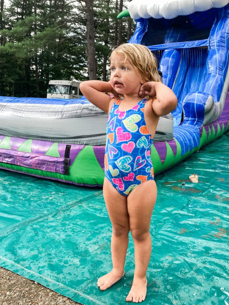 Young girl standing in front of a large inflatable water slide, wearing a colorful heart-print swimsuit with wet hair after water play at a luau-themed birthday party.