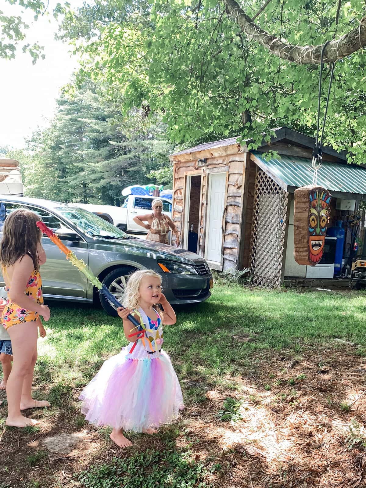 Young girl in a rainbow tulle skirt holding a bat and smiling as she prepares to hit a tiki piñata hanging from a tree, with other children and adults watching at a luau-themed outdoor party.