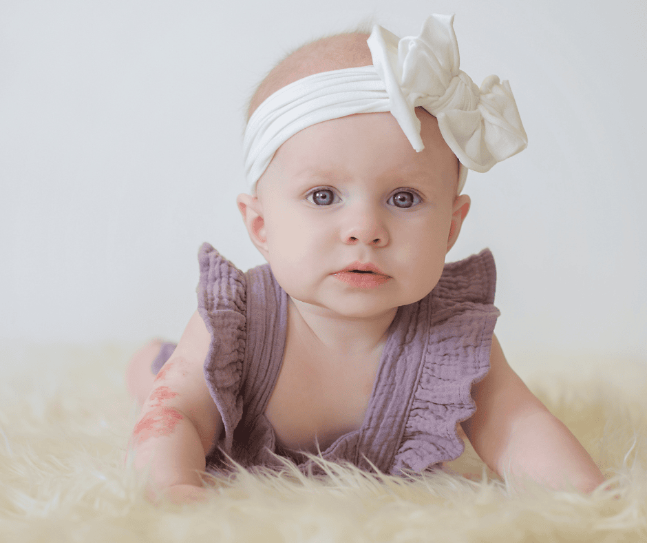 Baby girl wearing a white headband with a bow and a purple ruffle outfit, laying on a soft fuzzy blanket, looking at the camera.