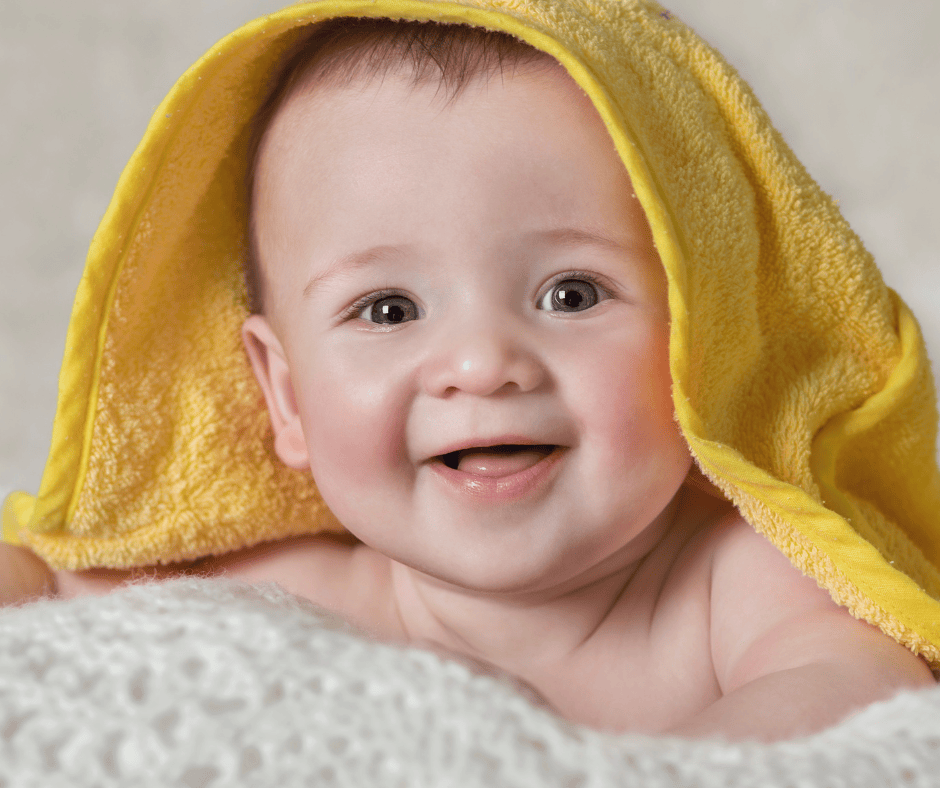 Smiling baby boy wrapped in a yellow towel, lying on a white blanket.