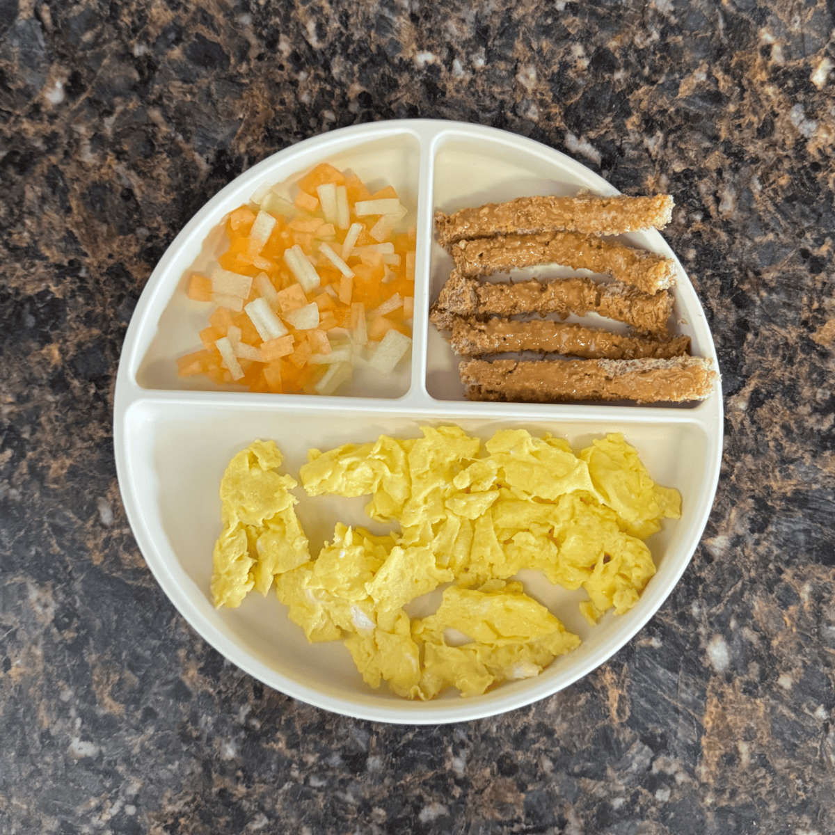 A toddler breakfast plate with three compartments: scrambled eggs, toast strips with peanut butter, and diced honeydew and cantaloupe.
