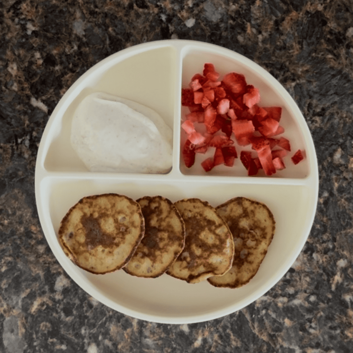 Toddler breakfast with banana and egg pancakes, diced strawberries, and a side of plain Greek yogurt with cinnamon — an easy high-protein meal for 1-year-olds.