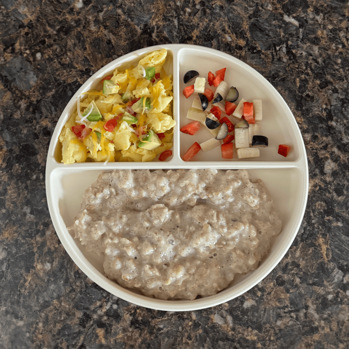 Toddler breakfast plate with maple cinnamon oatmeal, scrambled eggs topped with cheese, avocado, and tomato, and a side of diced strawberry, blueberry, and banana — a balanced breakfast idea for 12-month-olds.