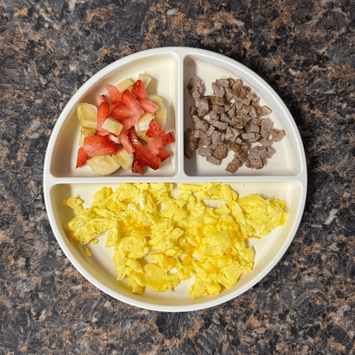 Toddler breakfast plate with scrambled eggs, diced breakfast sausage, and a mix of sliced banana and strawberry — a protein-rich and easy breakfast for 12-month-olds.
